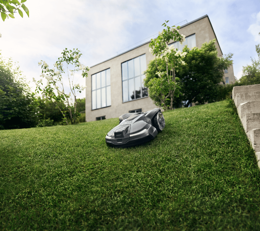 Automower working on downhill in a backyard with a house in the background